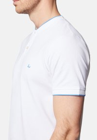 Biała koszulka polo z krótkim rękawem, z niebieskim wykończeniem kołnierzyka i listwą guzikową. Posiada haftowane logo na piersi. Wykonana z bawełny.