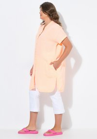 Maglietta lunga color pesco, con texture, chiusura a bottoni e tasche laterali, abbinata a pantaloni capri bianchi e infradito rosa.