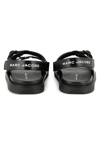 Mustat sandaalit säädettävillä remmeillä, joissa on valkoista "MARC JACOBS" -brändäystä, litteä pohja ja sileä nahkaverhoilu.