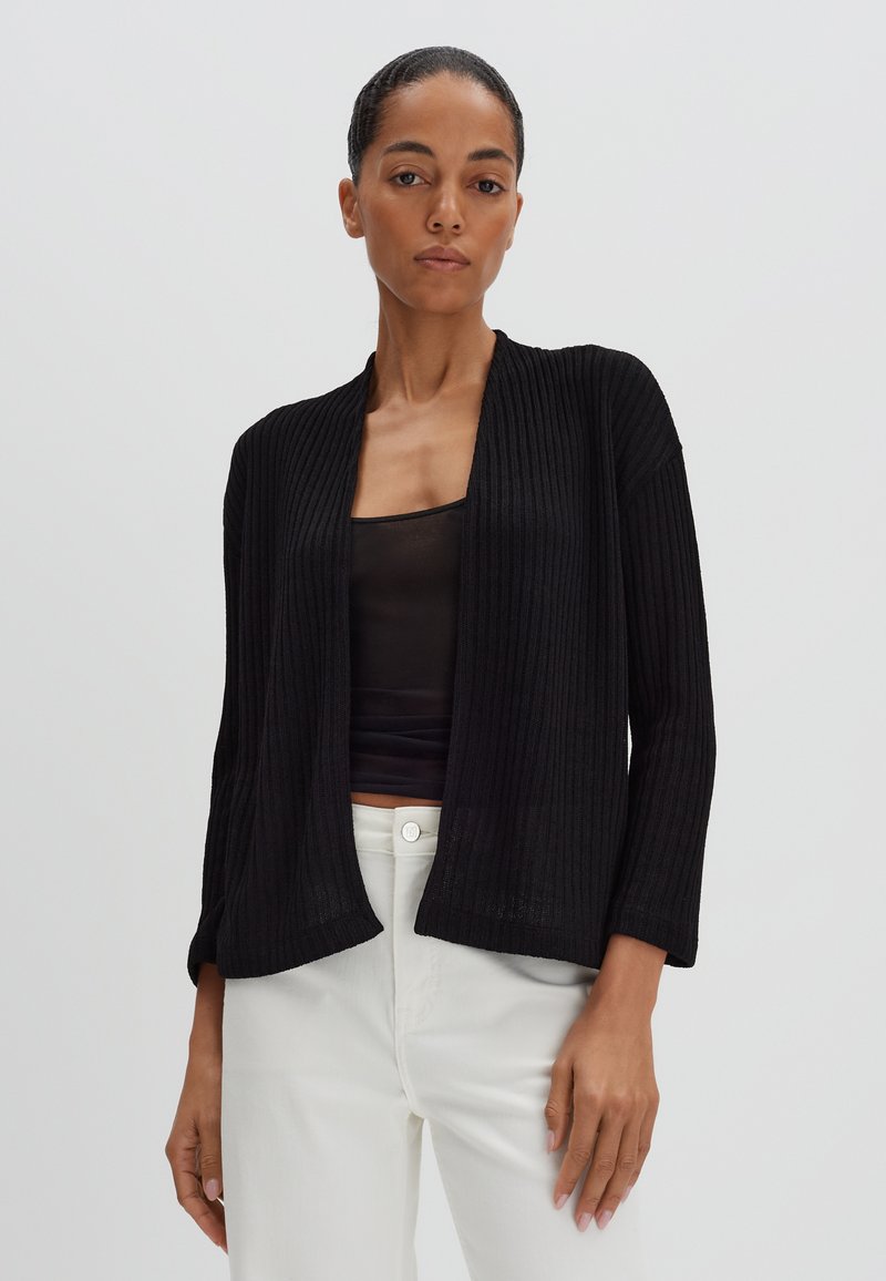Zwarte geribde cardigan met een open voorkant, lange mouwen en een relaxed fit, gedragen over een zwarte top en gecombineerd met witte broeken.