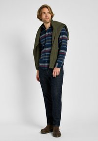 Camicia a righe in navy, rosso e verde indossata sotto un maglione lavorato in oliva. Jeans in denim scuro e stivaletti marroni alla caviglia. Modello in piedi in modo casual.