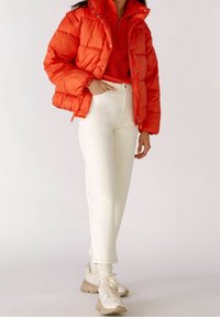 Veste matelassée rouge à fini brillant, col montant et boutons-pression, associée à un jean blanc taille haute et des baskets beige.