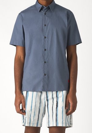 Mann trägt ein kurzärmeliges blaues Hemd mit Knöpfen und weiße Shorts mit vertikalen blauen und schwarzen Streifen vor einem schlichten Hintergrund.