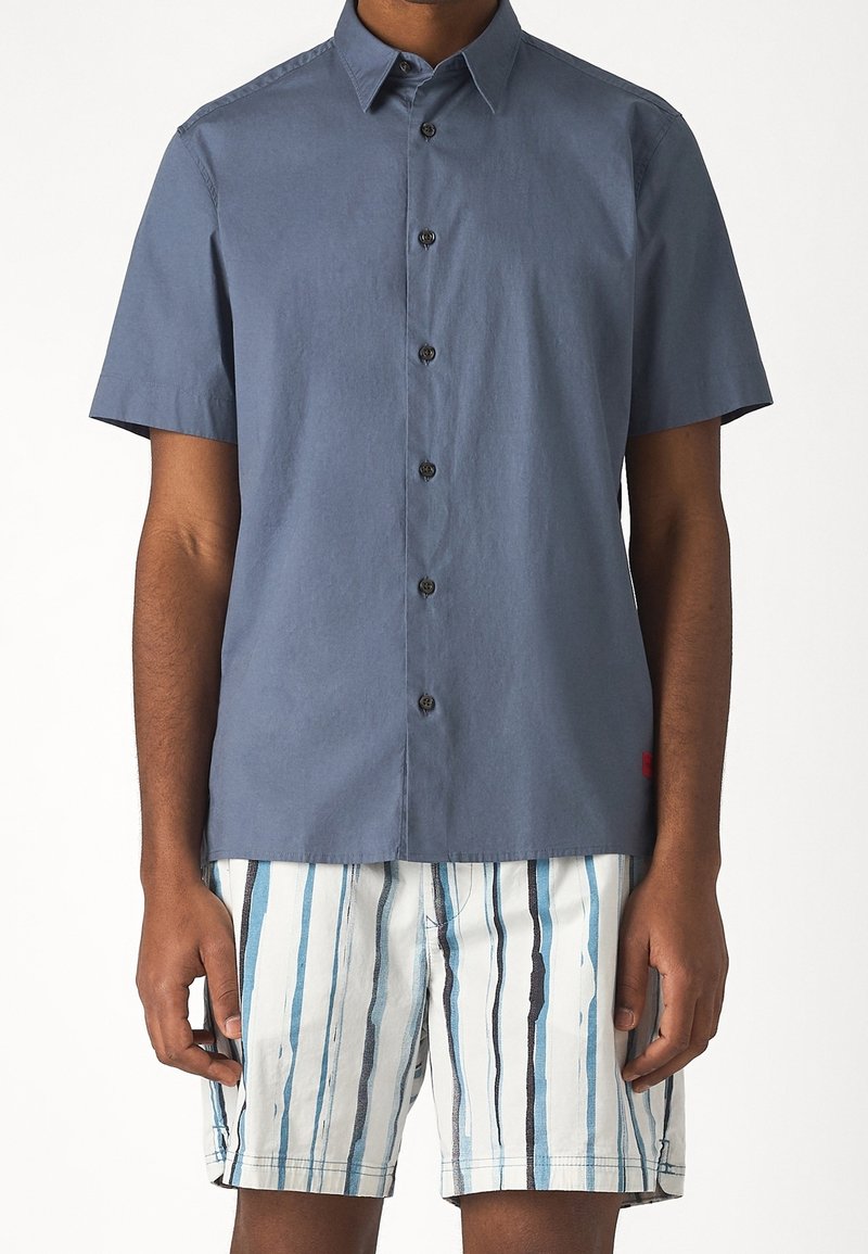 Homme portant une chemise bleue à manches courtes avec boutons et un short blanc à rayures verticales bleues et noires sur un fond uni.