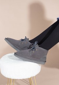 Stivaletti grigi in suede con lacci, dotati di occhielli in ottone, suola testurizzata e cuciture visibili, esposti su uno sgabello bianco in peluche.