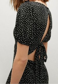 Czarna sukienka w białe kropki, z bufkowymi rękawami i odkrytymi plecami związanymi kokardą. Lekki materiał o gładkiej fakturze.