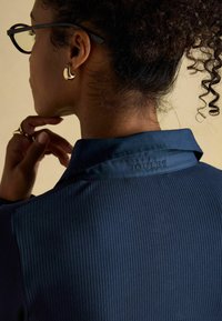 Navy blue collared shirt with ribbed texture, featuring embossed "JOULES" logo on the collar, worn by a person with curly hair and glasses.