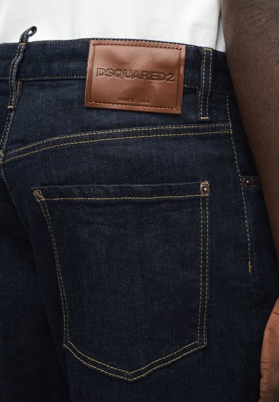 Σκούρα μπλε τζιν με κίτρινα ραφές, νιπ επάνω στην πίσω τσέπη και καφέ δερμάτινο ταμπελάκι DSQUARED2 πάνω από την τσέπη, φορεμένα με λευκό πουκάμισο.