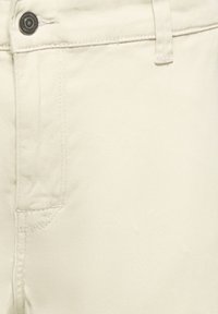 Pantalon beige en mélange de coton avec une texture lisse, doté d'une poche avant et d'une fermeture par bouton métallique, avec des détails de couture visibles.