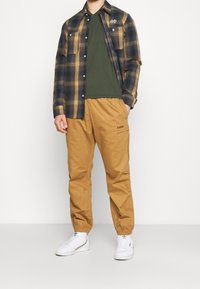 Károvaná flanelová košile v námořnické modři a hořčicové, zelené tričko s výstřihem do V, béžové kalhoty s elastickým pasem, bílé tenisky s akcenty a viditelným logem.