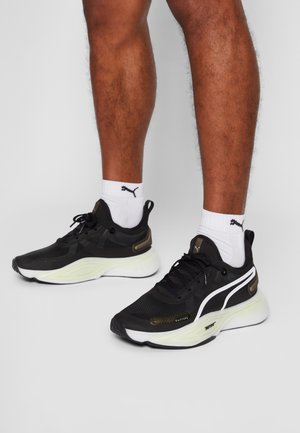 Schwarze Sportschuhe mit einem strukturierten Mesh-Obermaterial, weißer Sohle und goldenen Akzentdetails. Getragen mit weißen Socken, die ein Logo zeigen.