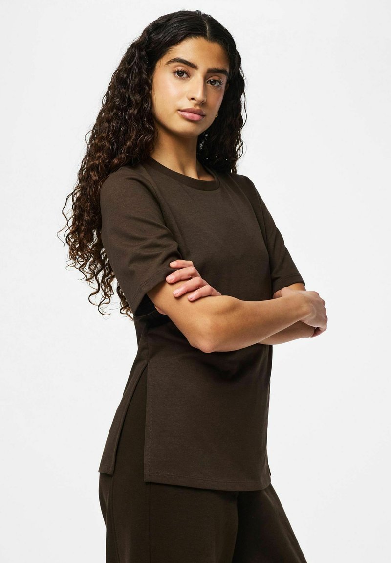 Femme aux longs cheveux bouclés et foncés, vêtue d'une chemise à manches courtes marron foncé et d'un pantalon assorti, debout les bras croisés devant un fond uni.