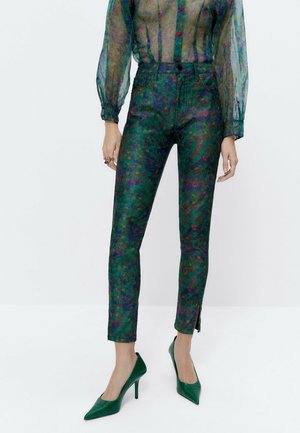 Mujer con blusa floral transparente, pantalones pitillo verdes con cintura alta y aberturas en los tobillos, y zapatos de tacón verdes con punta puntiaguda.