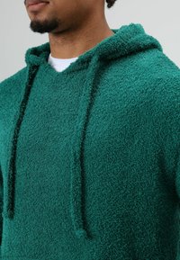 Sweat à capuche vert, duveteux, avec une finition texturée. Comprend une capuche et des cordons de serrage, avec un ajustement près du cou. Design simple, sans ornements.