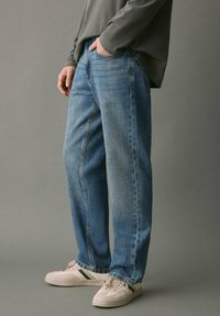 Svetlo modri jeans s sproščenim krojem, ki vključujejo klasičen dizajn s petimi žepi. Nosi se z dolgemi rokavi sivo majico in belimi supergami.