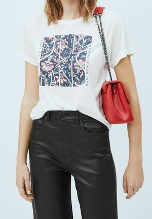 Vrouw die een wit T-shirt met een grafische print en bloemmotief draagt, zwarte leren broek, en een rode schoudertas met kettingriem bij zich heeft.