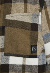 Braun-beige karierte Hemd mit einer Brusttasche, das ein minimalistisches Stoffetikett aufweist. Weicher Stoff, wolkenähnliches Material.