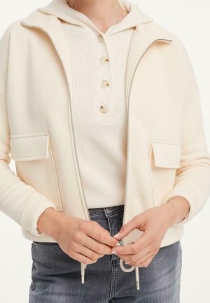 Person trägt eine cremefarbene Jacke mit Reißverschluss über einem gerippten, geknöpften Pullover und verschließt die Jacke, kombiniert mit grau gewaschenen Jeans.