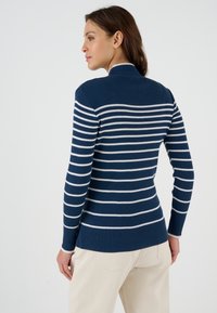 Vrouw die zijwaarts kijkt, gekleed in een marineblauw geribd sweater met witte horizontale strepen en lichtbeige broek tegen een effen achtergrond.