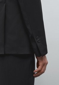Schwarze maßgeschneiderte Anzugjacke mit glatter Textur, ausgestattet mit einem Schlitzausschnitt und einem vierknöpfigen Detail am Ärmel. Minimalistisches Design.