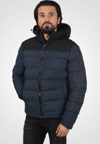 Nave plava puffer jakna s crnim gornjim panelom, zatvaranjem na zip i kapuljačom. Sadrži kviltanu teksturu i rebraste manžete, u kombinaciji s tamnim trapericama.