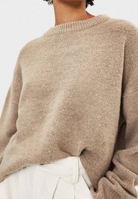 Person wearing a loose, textured beige sweater tucked into high-waisted off-white pants with pleats and belt loops.