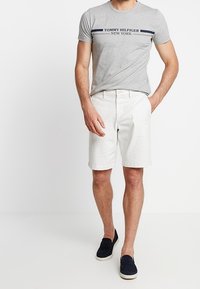 Graues Baumwoll-T-Shirt mit dem Logo "TOMMY HILFIGER NEW YORK", kombiniert mit weißen Shorts. Das Modell trägt marineblaue Slipper. Einfaches, lässiges Outfit.
