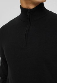 Černý zipový svetr s vysokým límcem, vyrobený z měkkého pleteného materiálu. Má čtvrtinový zip a hladkou texturu.