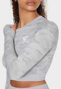 Grauer langärmeliger Cropped-Top mit einem Camouflage-Muster, rundem Ausschnitt und geripptem Saum. Verfügt über ein weißes Logo auf der Brust. Weiche Textur.