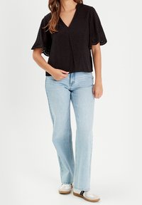 Blouse noire à manches courtes avec décolleté en V, présentant des motifs en points texturés et une coupe légèrement ample, associée à un jean flare bleu clair.