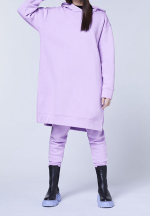 Persona con vestido sudadera lavanda extragrande con pantalones a juego y botas negras, de pie frente a un fondo liso.