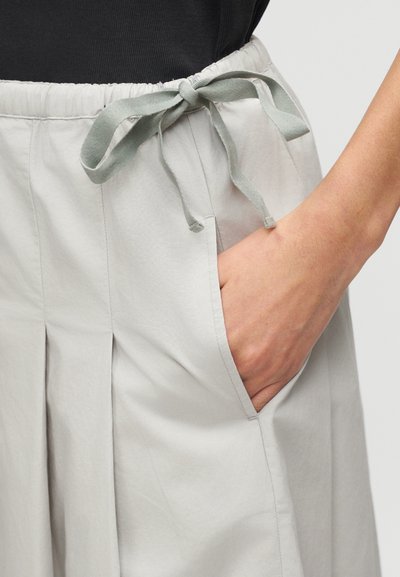 Φούστα ανοιχτό γκρι με πιέτες και υφασμάτινο φιόγκο στη μέση, χέρι τοποθετημένο στην πλαϊνή τσέπη, φορεμένη με μαύρο τοπ.