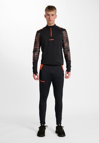 Czarny top sportowy z wzorzystymi rękawami, pomarańczowymi dodatkami, kołnierzem na zamek do ćwiartki, w połączeniu z czarnymi dopasowanymi spodniami z pomarańczowym logo.