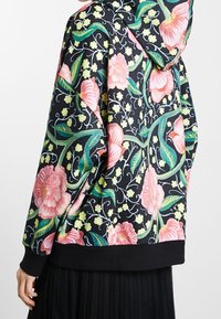 Kapuzensweatshirt aus schwarzem Stoff, verziert mit pinken Blumen und grünen Blättern, ausgestattet mit einem gerippten schwarzen Saum und weicher Textur.