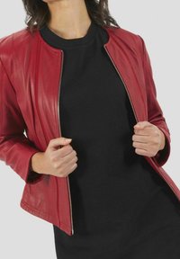 Veste en cuir rouge à coupe ajustée, fermeture éclair et surface légèrement texturée. Portée sur un haut noir, affichant des lignes simples et modernes.