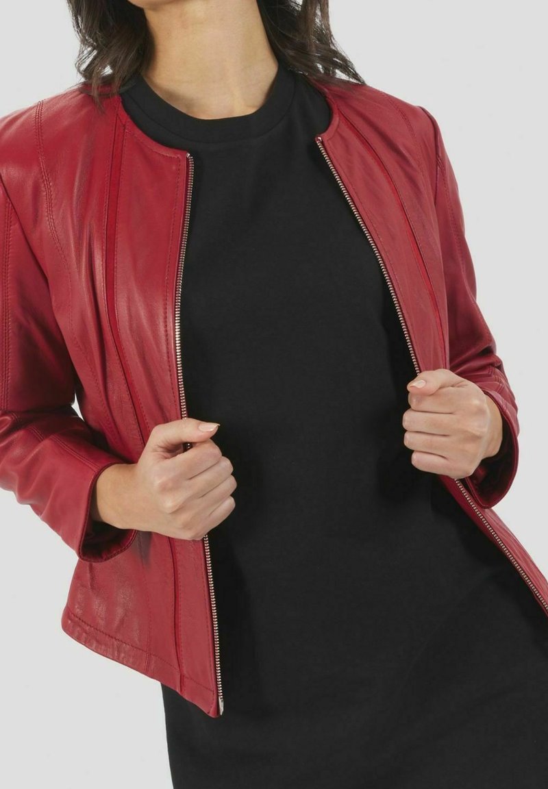 Veste en cuir rouge à coupe ajustée, fermeture éclair et surface légèrement texturée. Portée sur un haut noir, affichant des lignes simples et modernes.