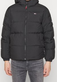 Černá prošívaná bunda s předním zipem, elastickými manžetami a kapucí. Design obsahuje horizontální prošití a malé logo na hrudi.