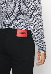 Uomo che indossa jeans neri con etichetta rossa "HUGO" in vita e una camicia a maniche lunghe bianco e nera a fantasia.