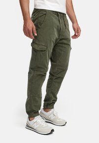 Pantaloni cargo verde oliva con polsini elastici, molteplici tasche e una vestibilità rilassata, realizzati in un tessuto morbido e testurizzato. Abbinati a sneakers grigie.