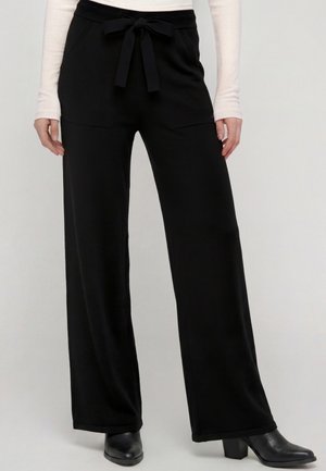 Pantaloni neri a gamba larga con cintura annodata davanti, indossati con stivaletti neri con tacco e top rosa chiaro a maniche lunghe.