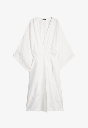 Biała sukienka o szerokich rękawach, z haftowanymi kwiatowymi detalami w formie wycięć oraz paskiem w talii na jednolitym tle.