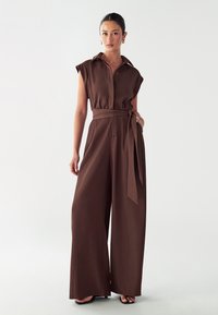 Bruin jumpsuit met korte mouwen, een kraag, knopen aan de voorkant en een tailleband. Heeft brede broekspijpen en een gladde stofstructuur.