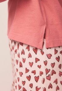Růžové bavlněné tričko s bočním rozparkem, doplněné světle růžovými pyžamovými kalhotami s rozptýlenými vzory červených srdcí. Hladká textura, ležérní design.