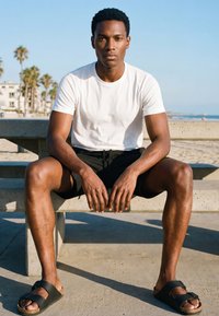 Fiatal férfi fehér pólóban, fekete rövidnadrágban és szandálban, aki egy betonpadon ül egy napsütötte tengerparton, háttérben pálmafákkal.