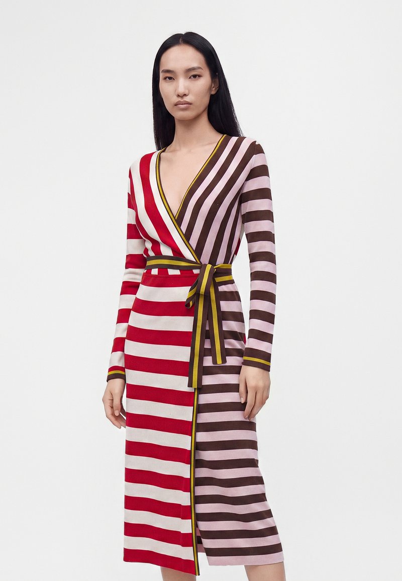 Femme portant une robe portefeuille à manches longues avec de larges rayures rouges et blanches d'un côté et des rayures roses et marron de l'autre, attachée avec une ceinture.