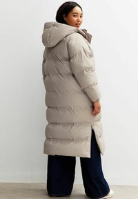Mujer con un abrigo acolchado largo beige con capucha, de pie de lado, combinado con pantalones oscuros de pierna ancha y zapatos blancos sobre un fondo liso.