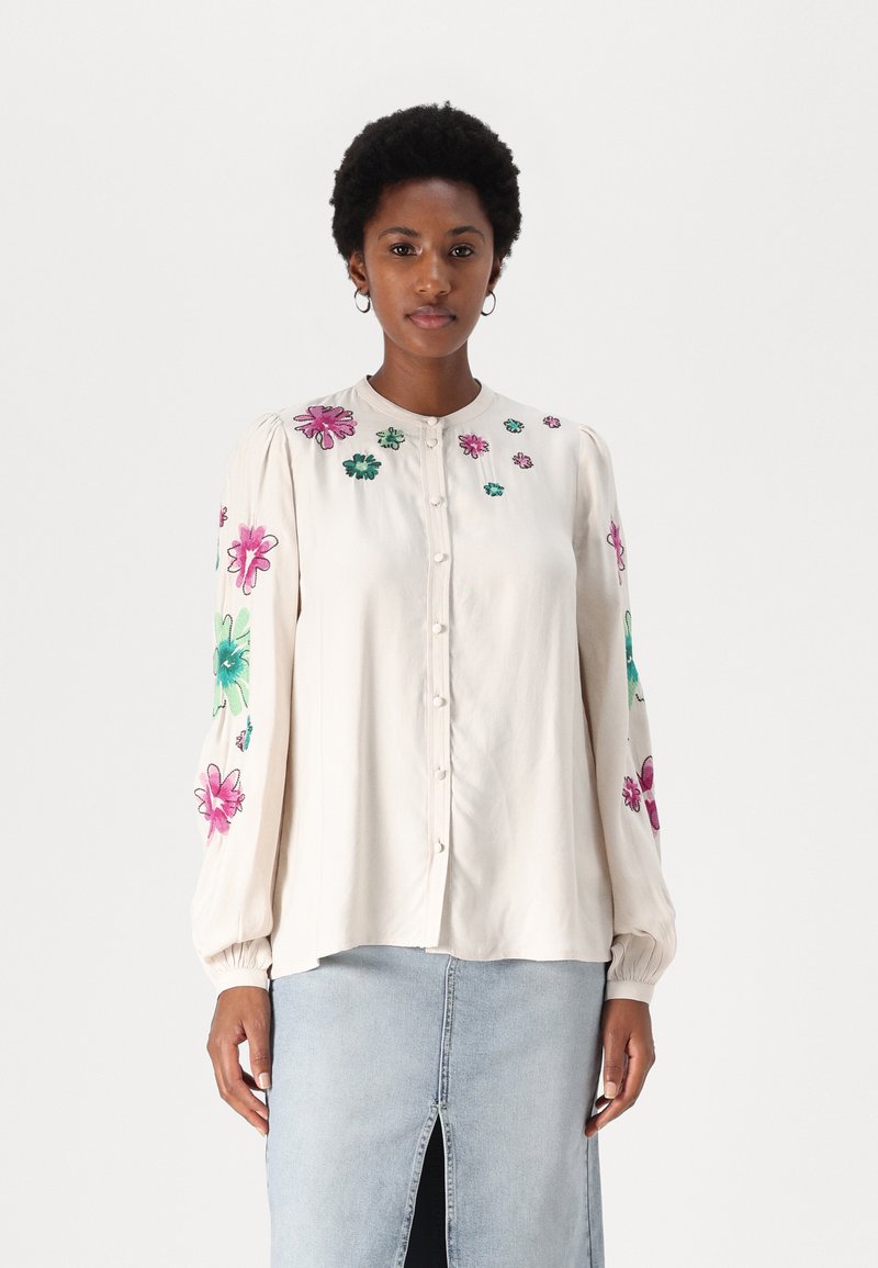Kremfarget skjorte med fargerik blomsterbroderi på ermene, med knappestolpe foran og myk, lett stoff. Kombinert med en denimskjørt.