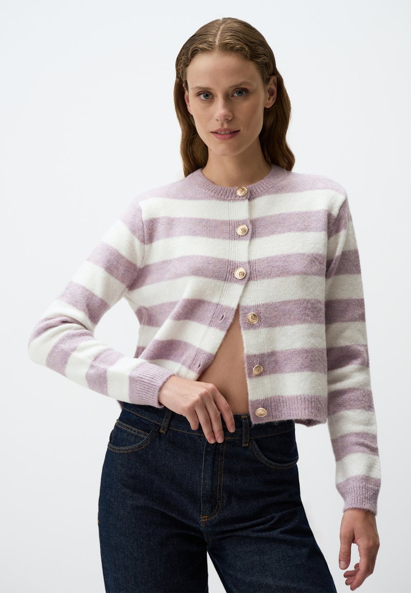 Cardigan a righe lilla e bianche con scollatura rotonda, sette bottoni e polsini a coste. Indossato su jeans denim blu scuro.