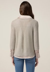 Strikket sweater i lys beige med et struktureret mønster, der har ærmer til albuen og et V-formet rygdesign over en kravebluse.