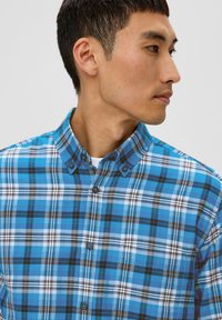 Chemise à boutons en motif tartan bleu comportant des nuances de bleu, noir et blanc. Fabriquée en tissu léger avec un col et des détails de boutons.